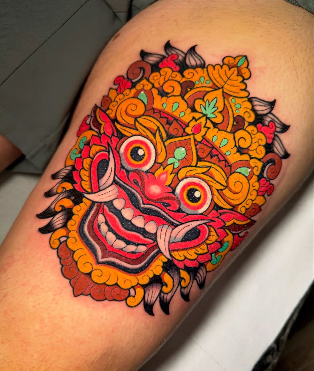 Colorful mask-like face tattoo featuring large red eyes, ornate orange and red patterns, decorative scrolls, and green leaf accents on the upper arm.