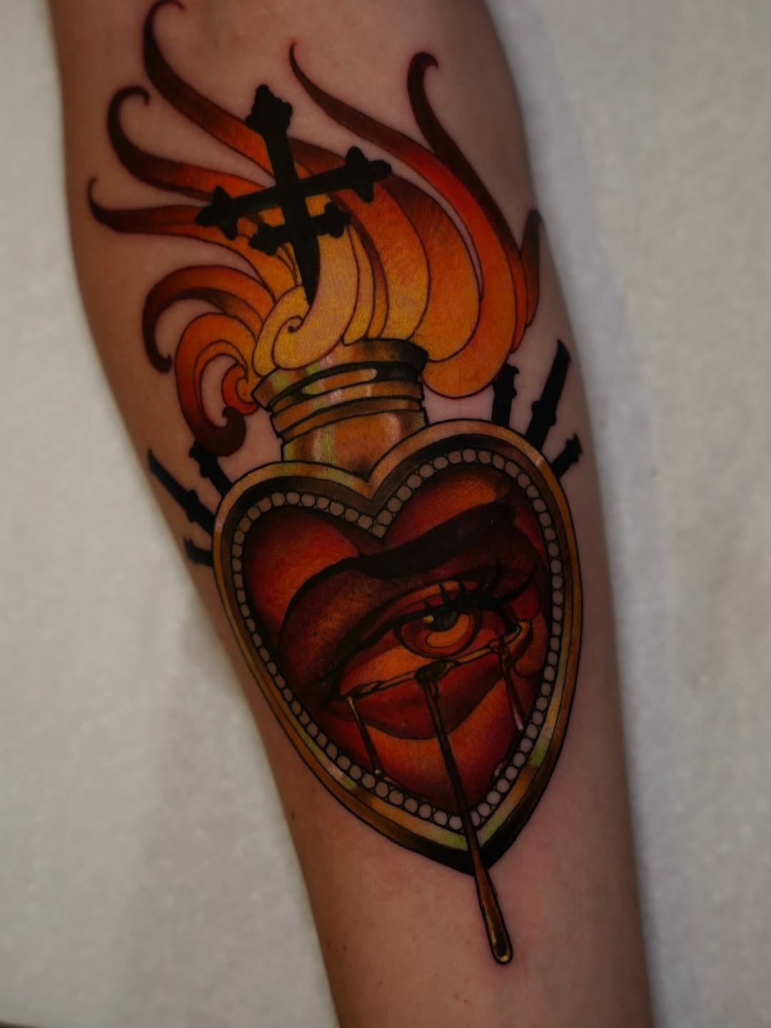 Sacred heart tattoo with flaming torch on upper portion, detailed anatomical heart with cross overlay, ornamental beading, and black ribbon, rendered in red, orange, and yellow on inner arm.