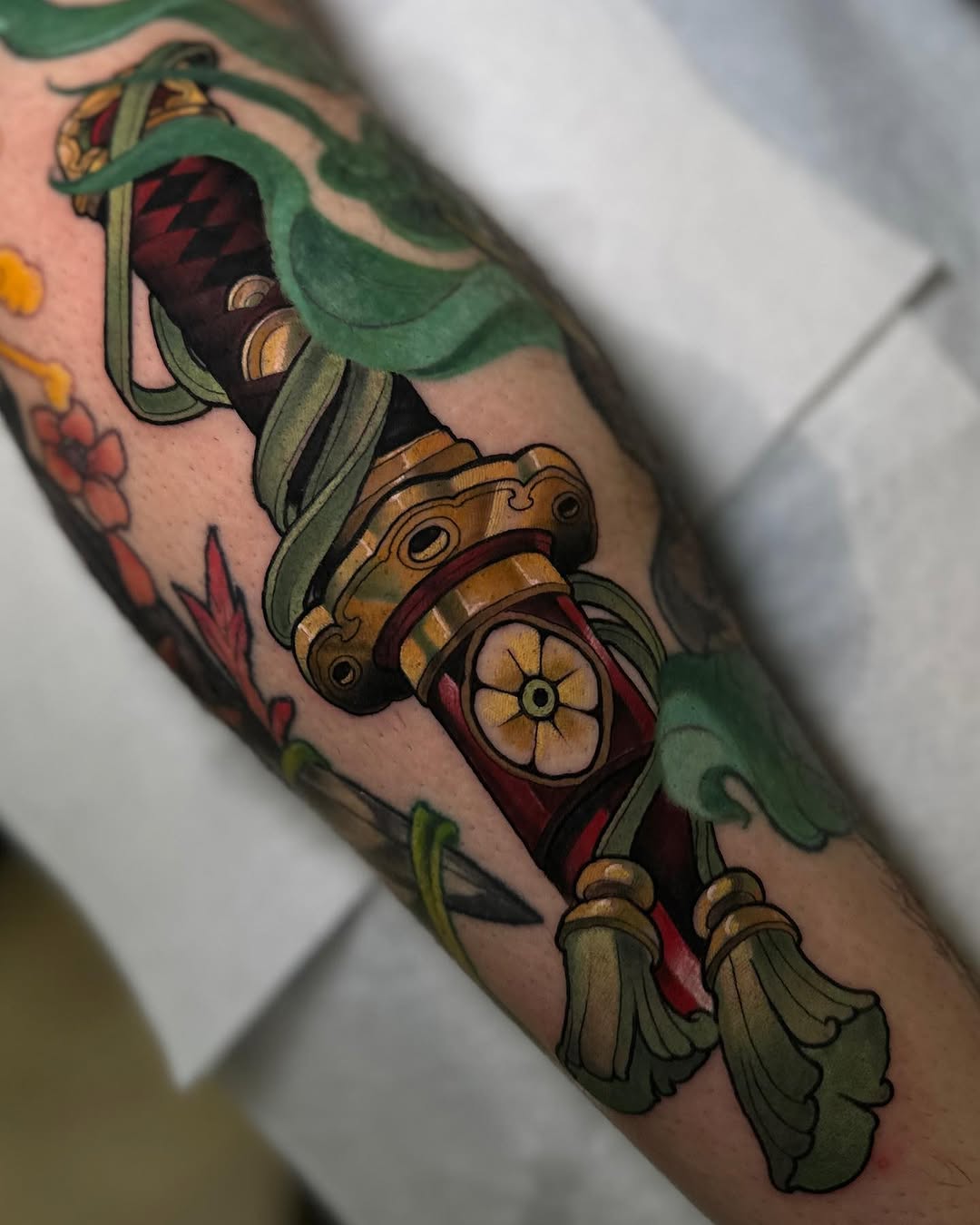 Colorful tattoo on an arm featuring an ornate mechanical device with green ribbons, gold gears, and red accents, accompanied by floral elements.