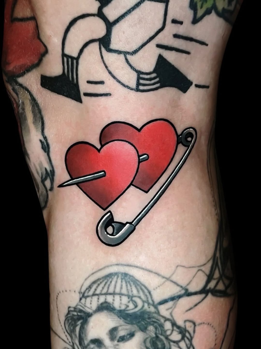 Two red hearts pierced by a needle and safety pin on the upper arm, with black line work and additional tattoos visible above and below.