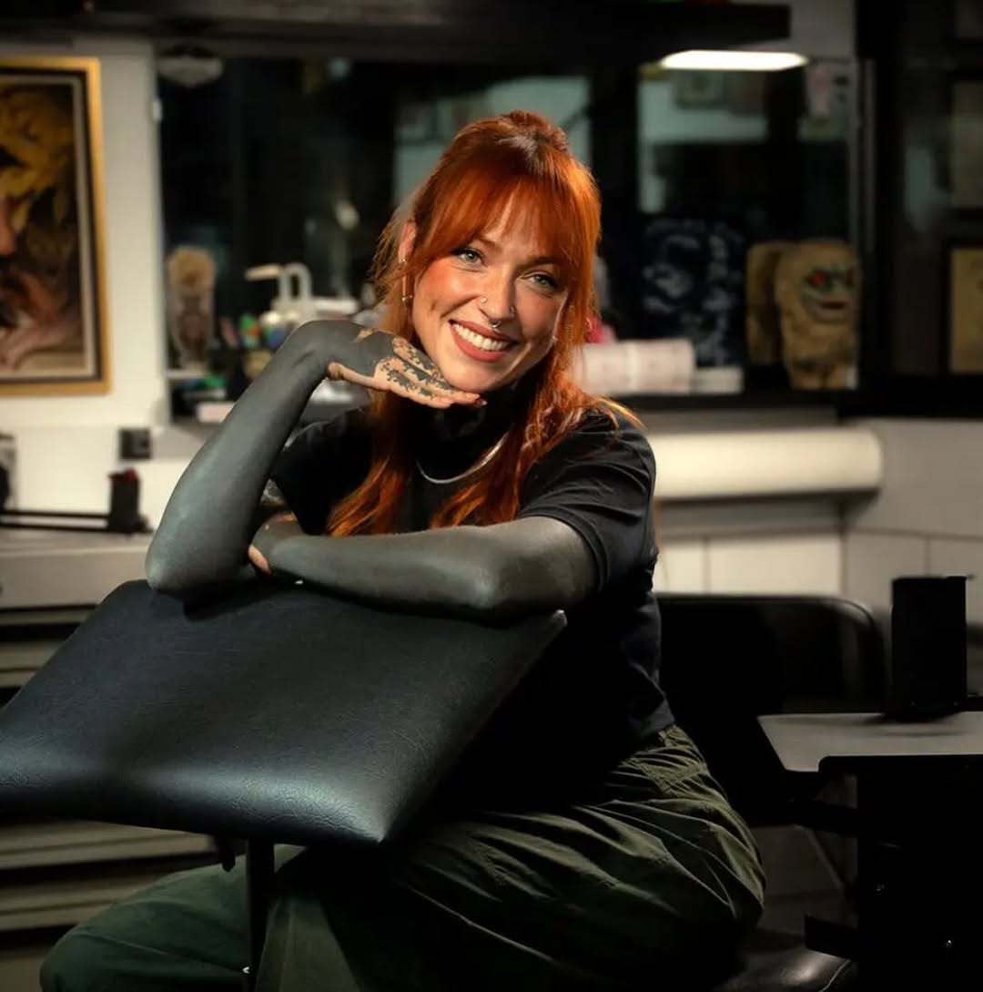 A woman with red hair and a neck tattoo sits in a tattoo studio, smiling at the camera while resting her arm on a black chair.