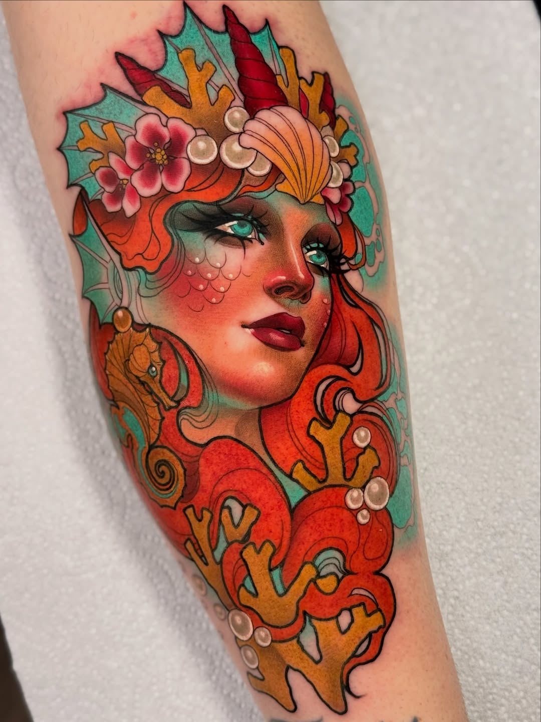 Colorful portrait tattoo of a woman's face with turquoise eyes, red hair, coral skin tones, and an elaborate headdress featuring flowers, pearls, and ornamental details on the forearm.