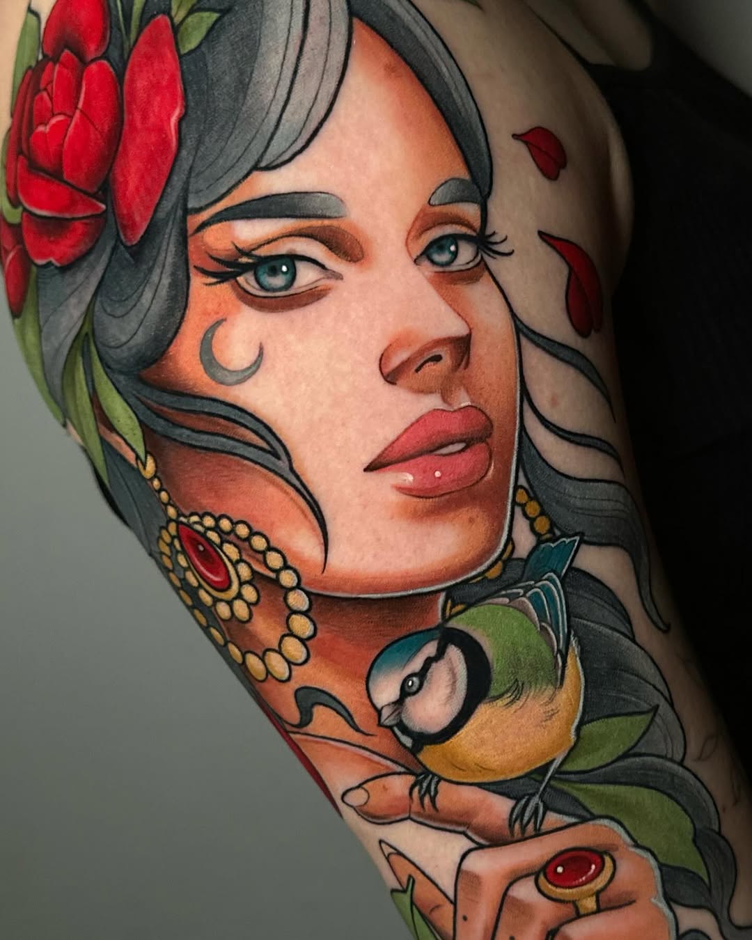 Colorful portrait tattoo on arm featuring a woman with dark hair, red flowers, a small bird, crescent moon marking, gold beaded necklace, and red petals.