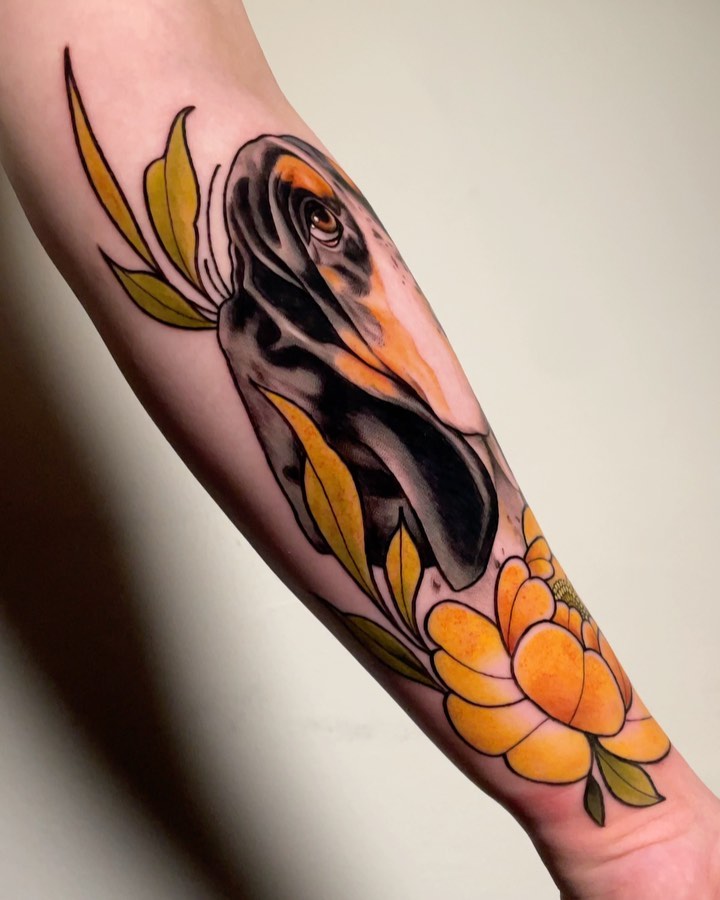 A bird's head in black and orange tones with yellow flowers below, tattooed on the inner forearm.