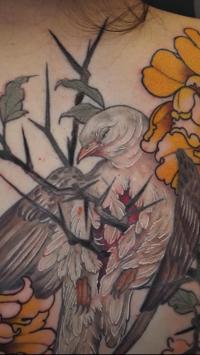 Shoulder tattoo featuring a detailed bird with spread wings, bare branches, and yellow flowers in orange and gray tones.