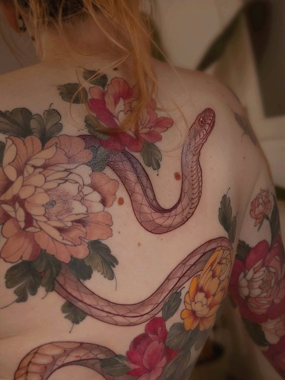 Colorful floral and serpent tattoo covering the shoulder and upper back, featuring peonies, roses, and intertwining snakes in red, pink, yellow, and green tones.
