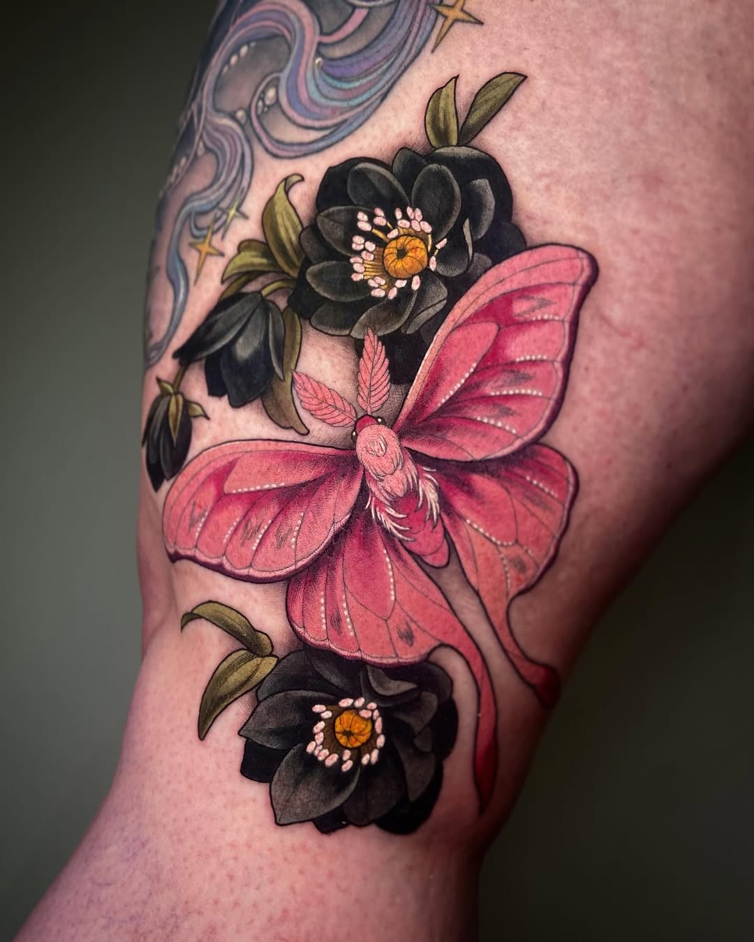 Pink butterfly with black flowers featuring orange centers and green leaves tattooed on upper arm, with colorful abstract design visible on shoulder.
