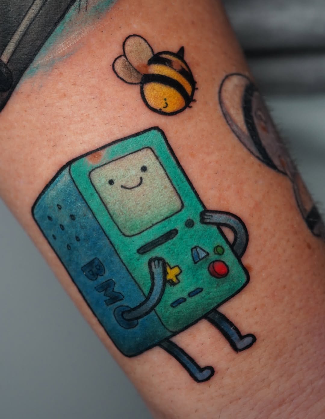 Colorful tattoo on arm featuring a smiling handheld gaming device in teal and green with a bee above it in yellow and black.