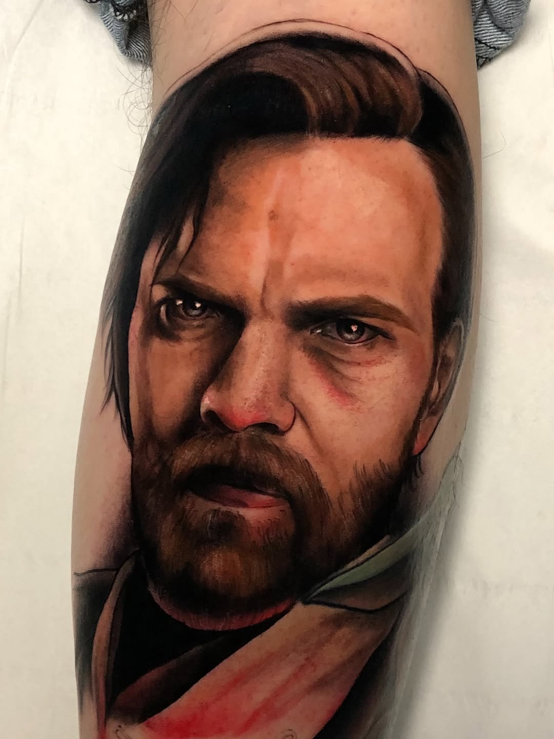 Portrait tattoo on inner arm of bearded man with long dark hair, rendered in brown, red, and flesh tones with detailed facial features and shoulder drapery.