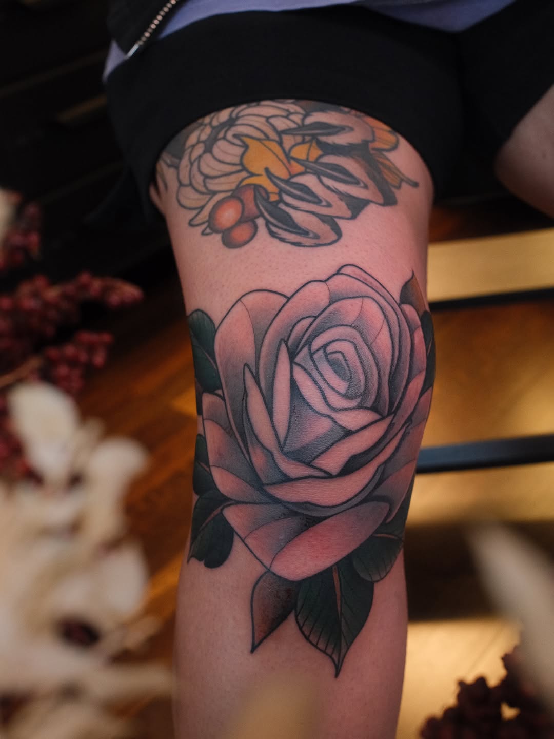 Rose tattoo with shaded petals and leaves on upper arm, with a bee design visible above on the same limb.