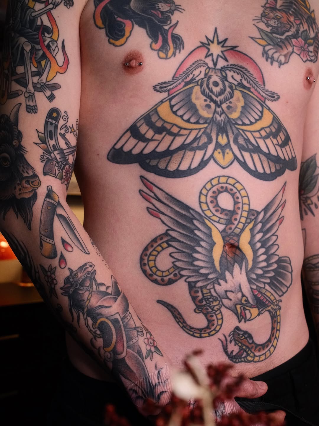 Chest tattoo featuring a large moth with spread wings and a star above, paired with a snake and ornamental circles below, rendered in black ink with gold and red accents.