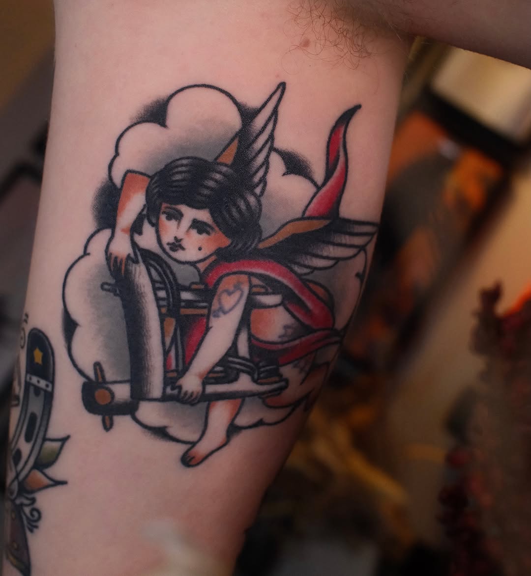 Winged devil character with horns and red cape seated on clouds, tattooed on upper arm in black, red, and orange ink.