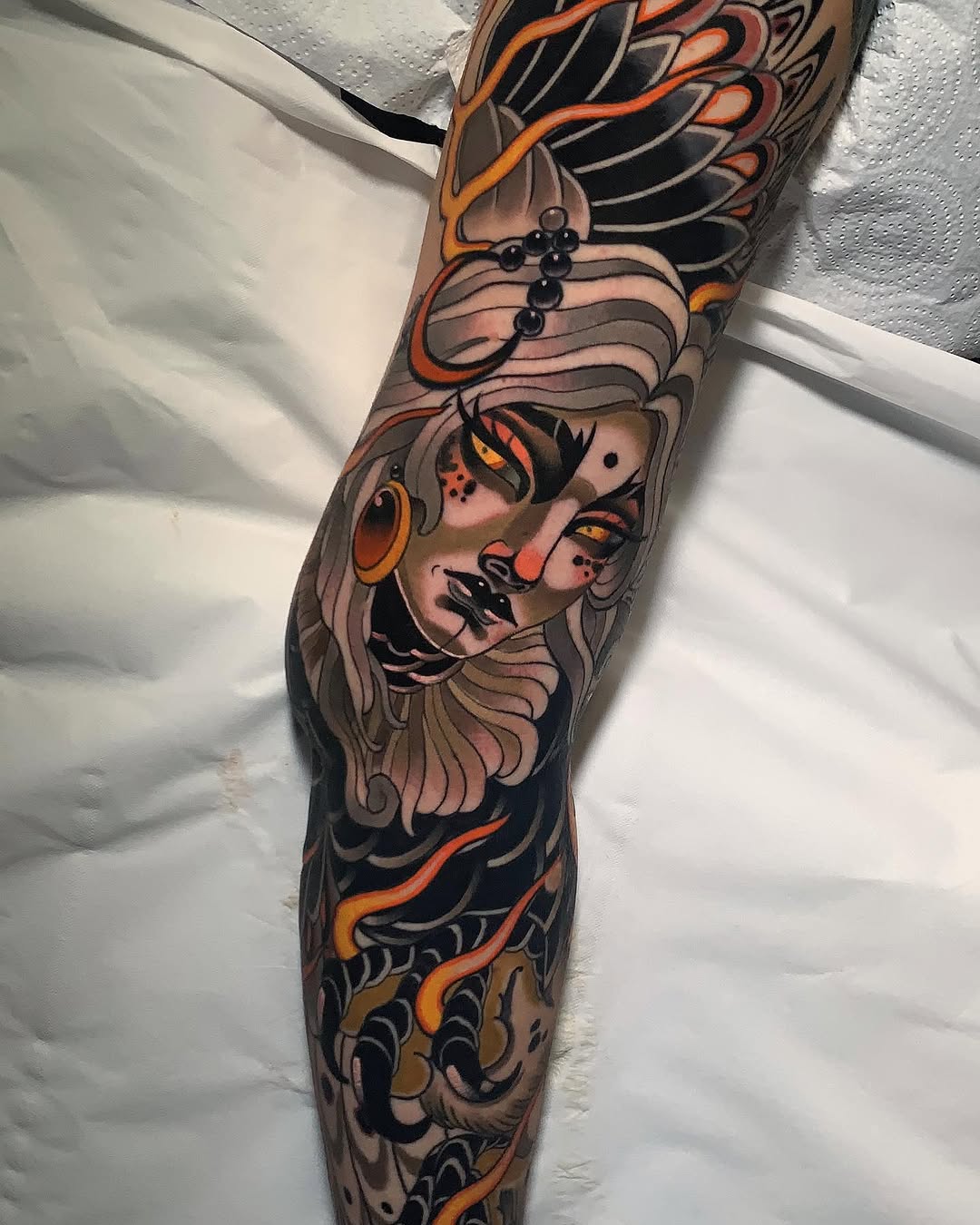 Full leg sleeve tattoo featuring a winged female portrait with cross motif, orange and black decorative elements, and ornamental patterns throughout.