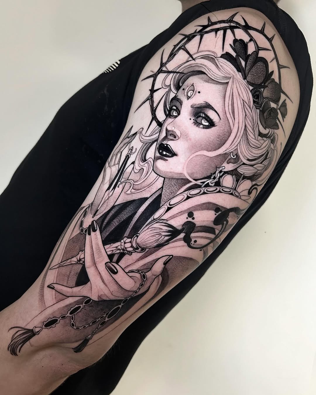 Portrait tattoo of a woman with long wavy hair, crown of thorns, roses, chains, and ornamental details on the upper arm in black ink with skin tone shading.