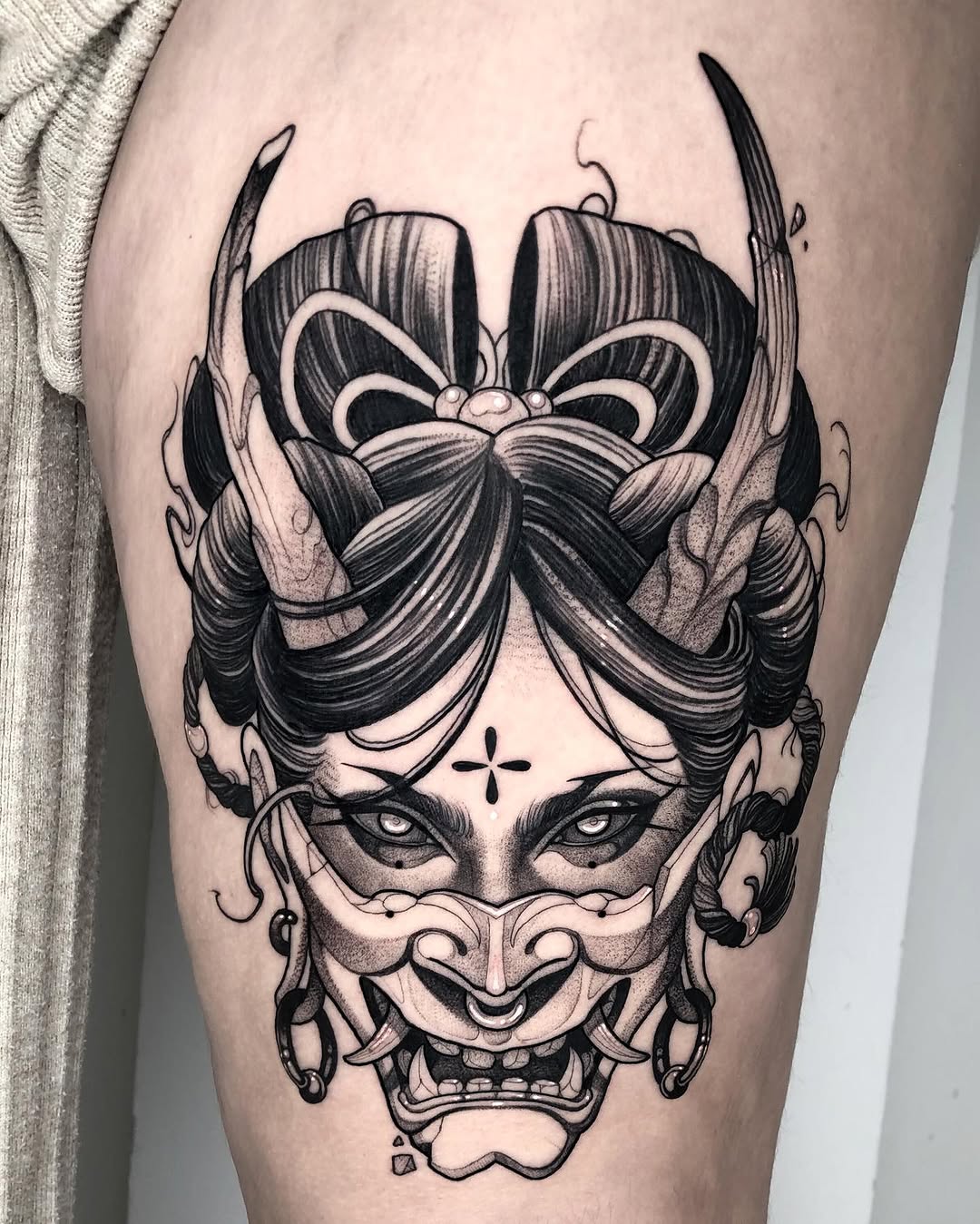 Detailed black ink tattoo on thigh featuring an ornate face with a large bow, mask markings, demon-like features, and decorative elements throughout.