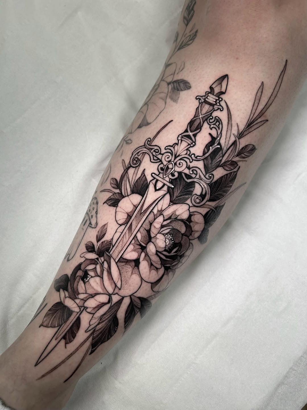 Ornate dagger with decorative scrollwork surrounded by large peonies and leafy botanicals on the forearm in black ink.
