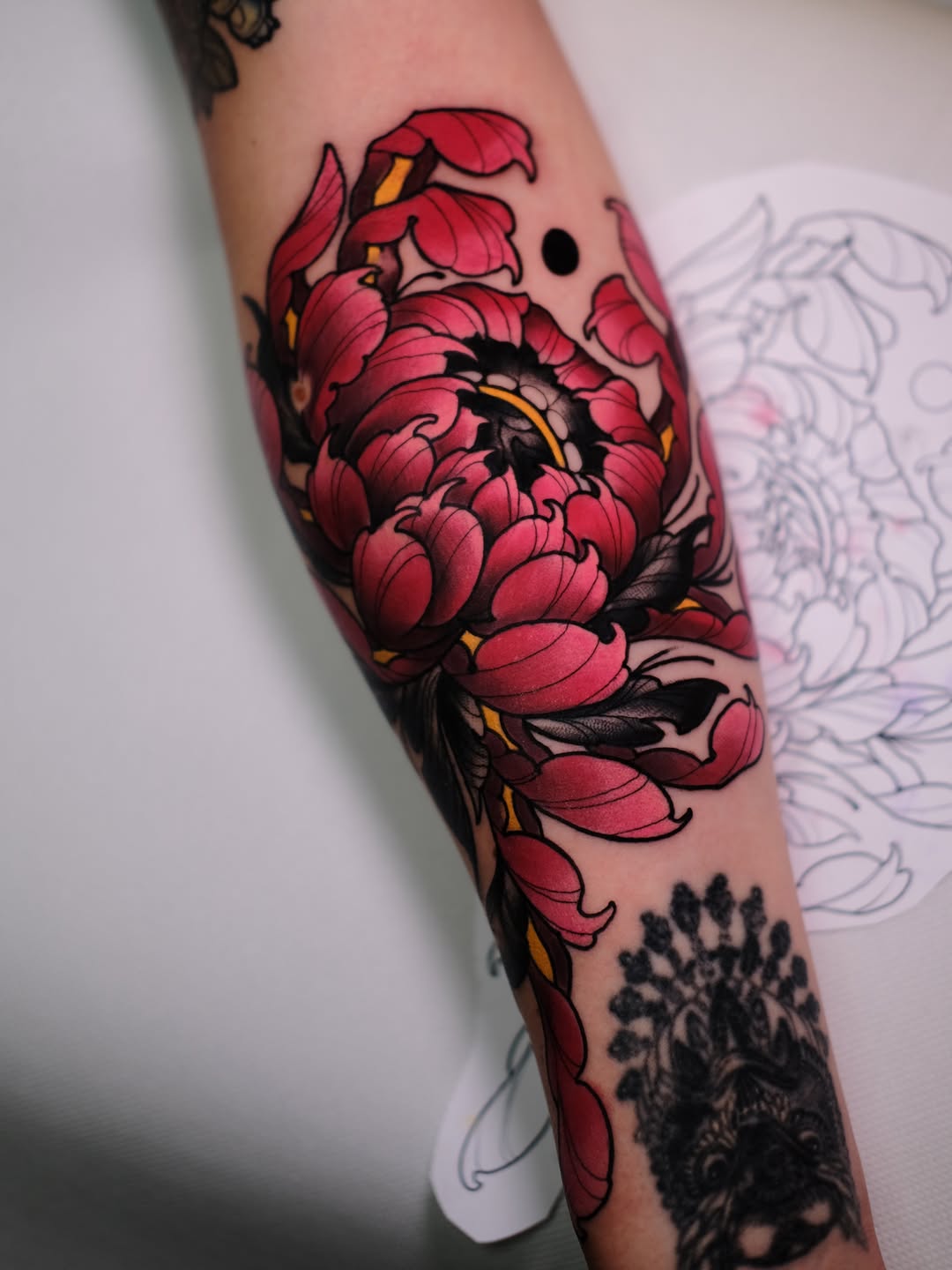 Red peony flower with black outlines and yellow accents tattooed on an arm, displayed next to a sketch reference.
