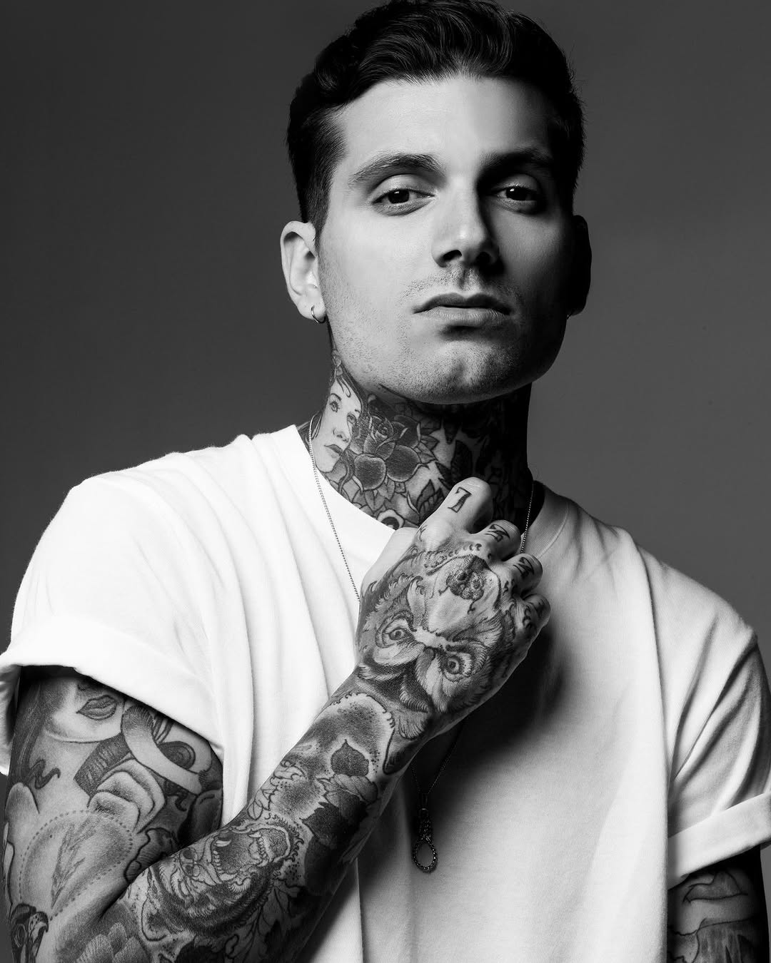 Man with extensive chest and arm tattoos featuring detailed floral and ornamental designs in black ink, wearing a white v-neck shirt.