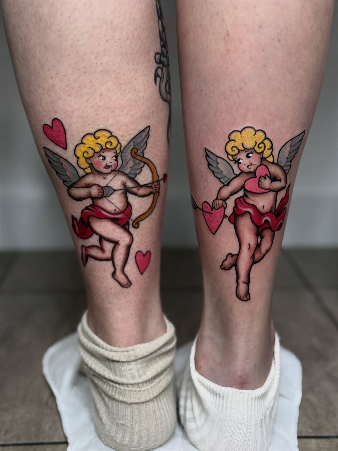 Matching cupid tattoos on both calves featuring winged cherubs with blonde curly hair, red hearts, and bows in pink, yellow, and red ink.
