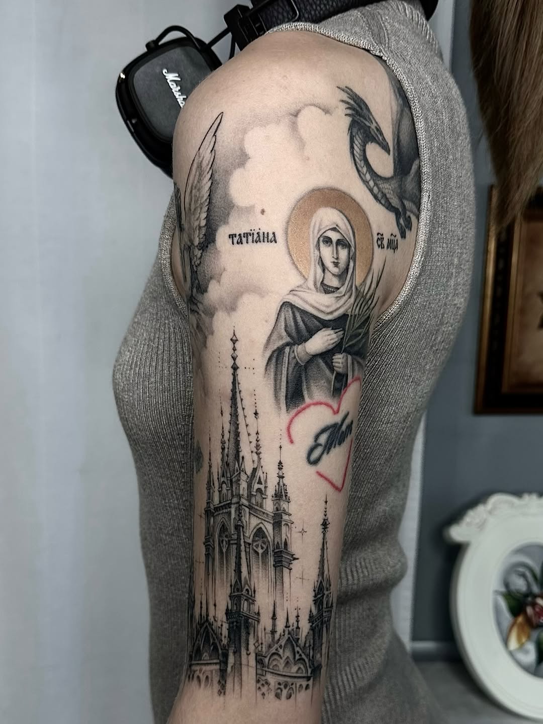 Religious-themed leg tattoo featuring a saint with halo, Gothic cathedral, birds, Cyrillic text, and red heart outline on the thigh.