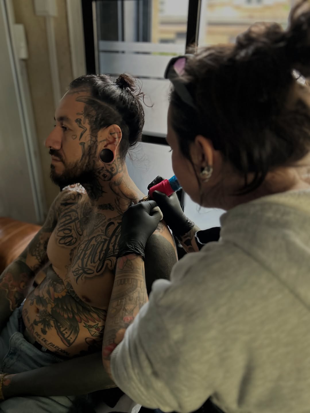 Tattoo artist applies red and black ink to client's shoulder and chest area during a studio session.