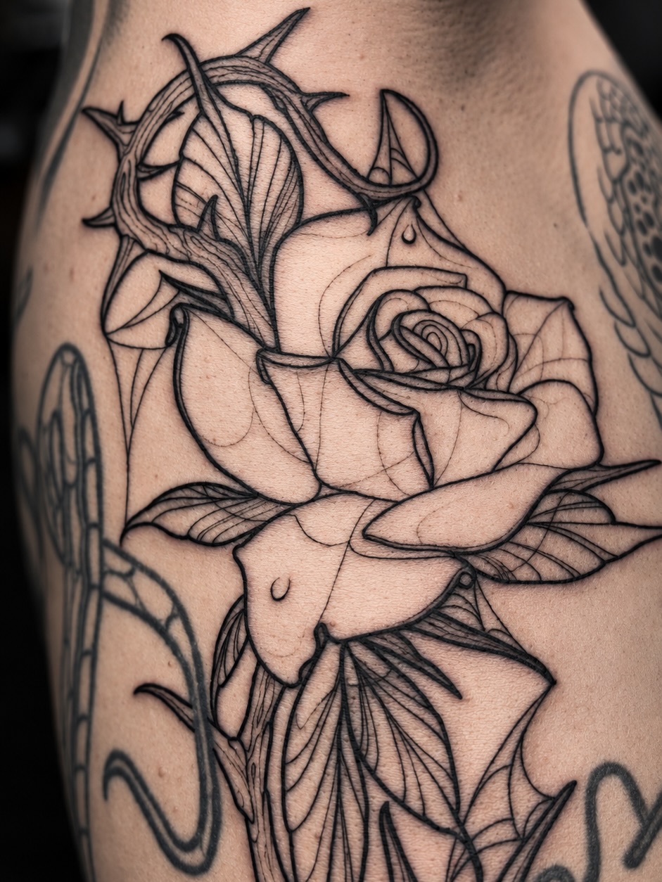 Black ink botanical tattoo on the upper arm featuring a rose with thorned stems and detailed leaf work with fine line details.