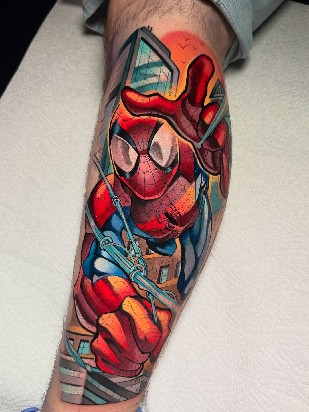 Spider-Man in dynamic pose with cityscape background tattooed on the forearm in vibrant reds, blues, oranges, and yellows.