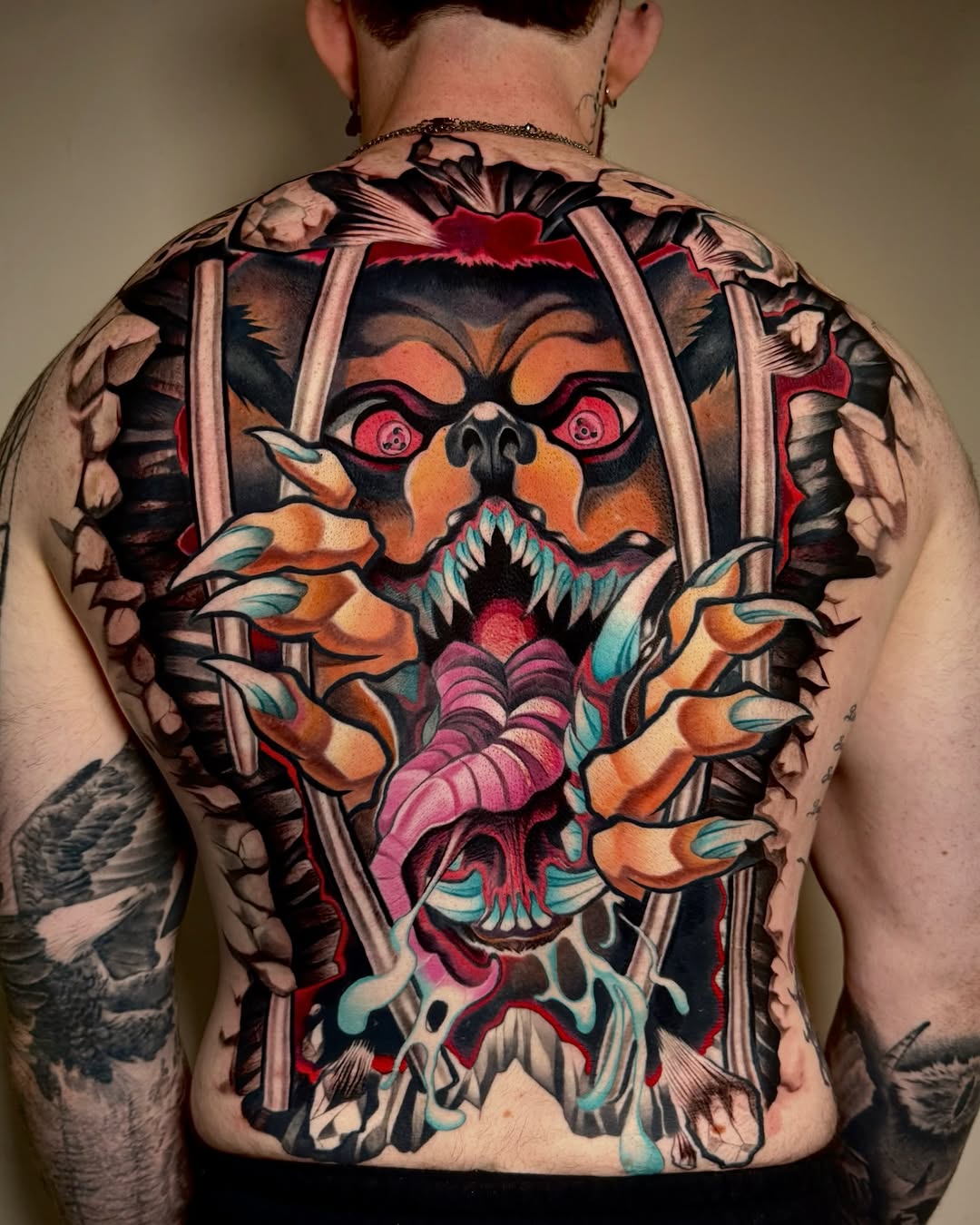 Large back tattoo featuring a demonic face with orange and black coloring, sharp teeth, pink tongue, and skeletal elements surrounded by ornamental details and flames.