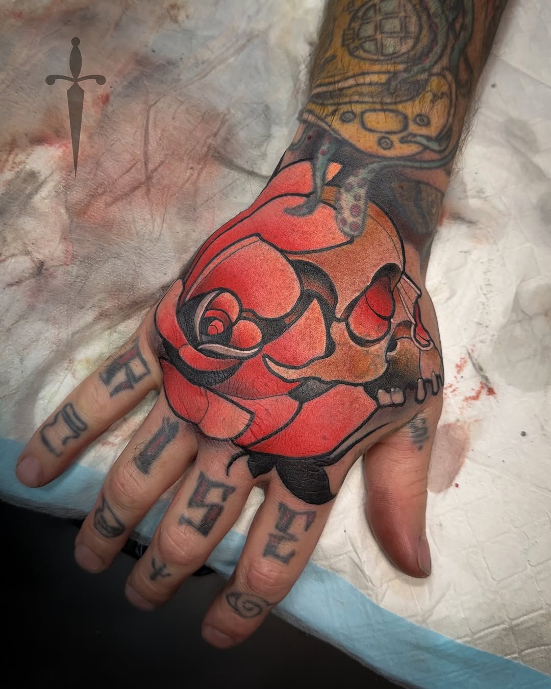Red and black skull with roses and black outline details tattooed on the back of a hand.