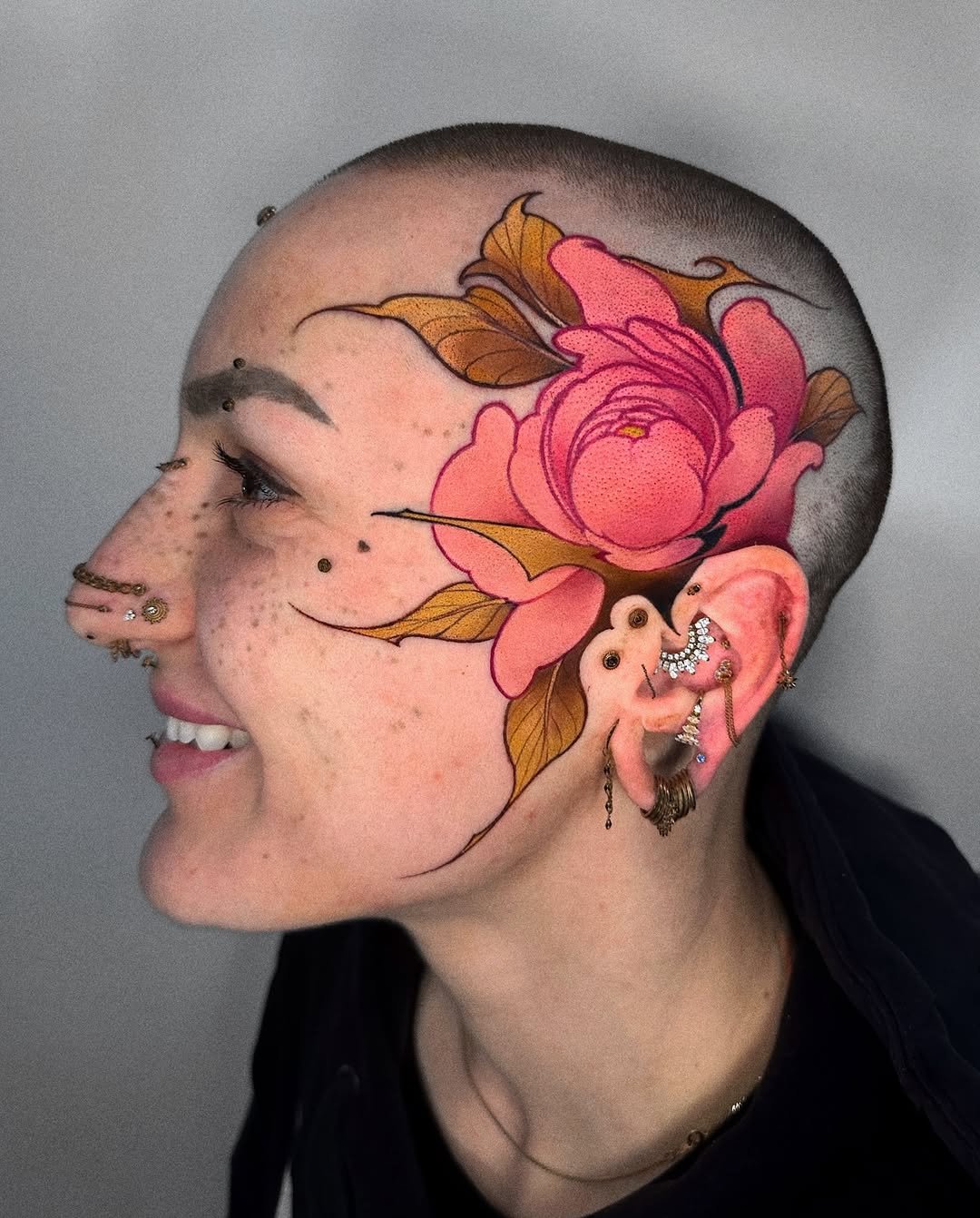 Side profile of a person's shaved head featuring a large pink and coral peony flower with golden-brown leaves extending across the scalp.