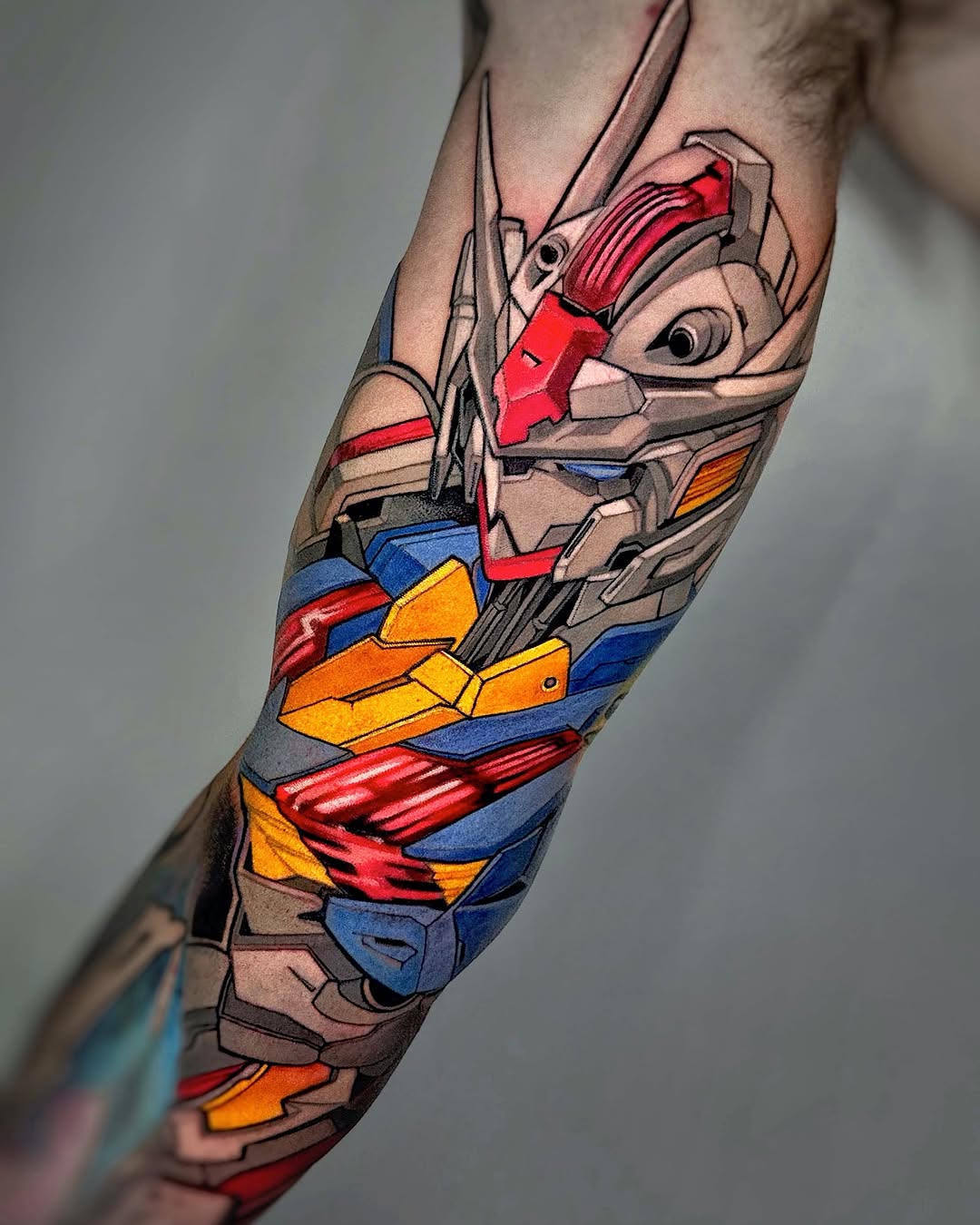 Colorful robot head tattoo on forearm featuring red, blue, yellow, and orange mechanical parts with gray outlines and details.