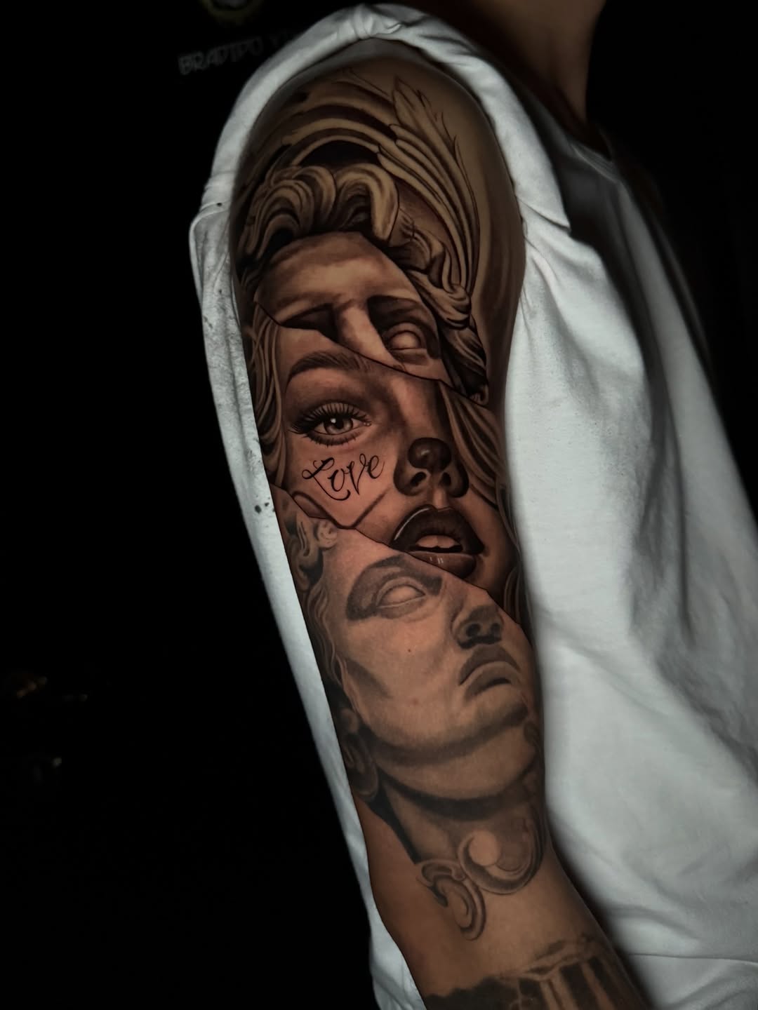 Detailed portrait tattoo on upper arm featuring two faces with ornamental flourishes and the word "Love" in script, rendered in brown and black ink.
