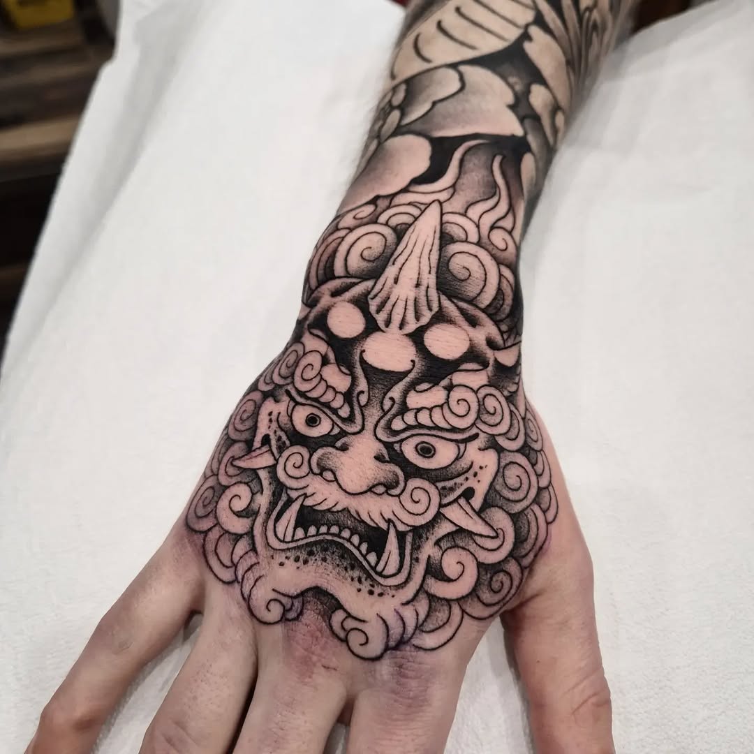 Ornate mythical beast head with scrolling flourishes and shell motifs tattooed on hand and forearm in black and grey ink.