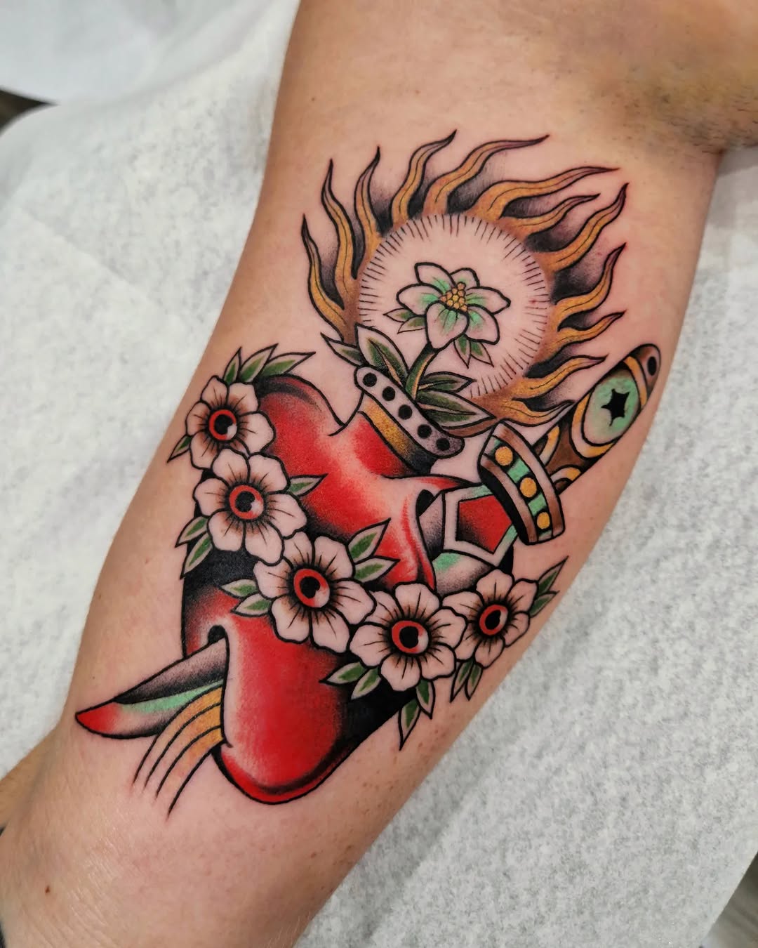 Sacred heart tattoo with flaming sun head, pink flowers, crescent moon, and star on inner forearm in red, black, gold, and green ink.