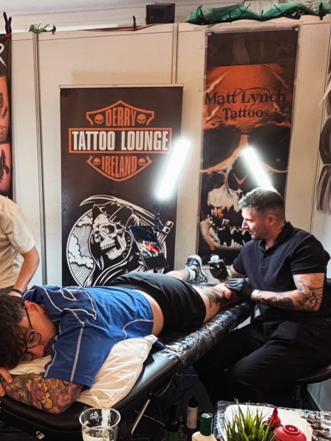 Tattoo artist applies a large skull and wing design on a client's leg at a convention booth.