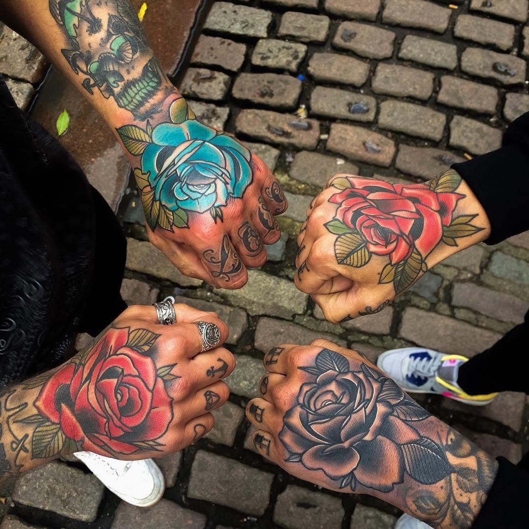 Four people display colorful rose tattoos on their hands and knuckles in red, blue, black, and green ink against a brick background.