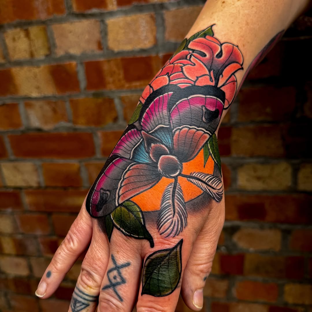 Colorful hand tattoo featuring two butterflies with red, pink, orange, and blue wings, green leaves, and black outlines on skin against a brick wall.