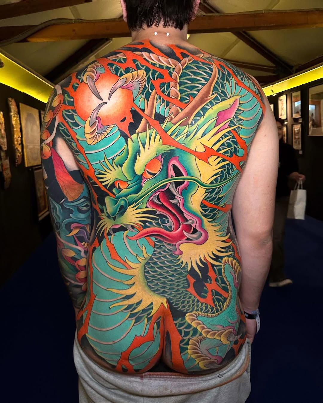 Colorful full back tattoo featuring dragons, flowers, and ornamental scales in red, teal, yellow, and pink on a shirtless person.