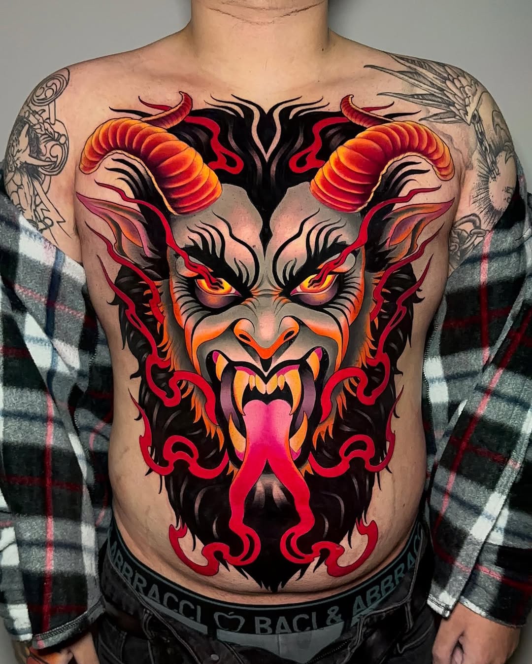 Vibrant demon face chest tattoo featuring red, orange, and black colors with curved horns, glowing eyes, and ornamental flourishes on a shirtless male torso.