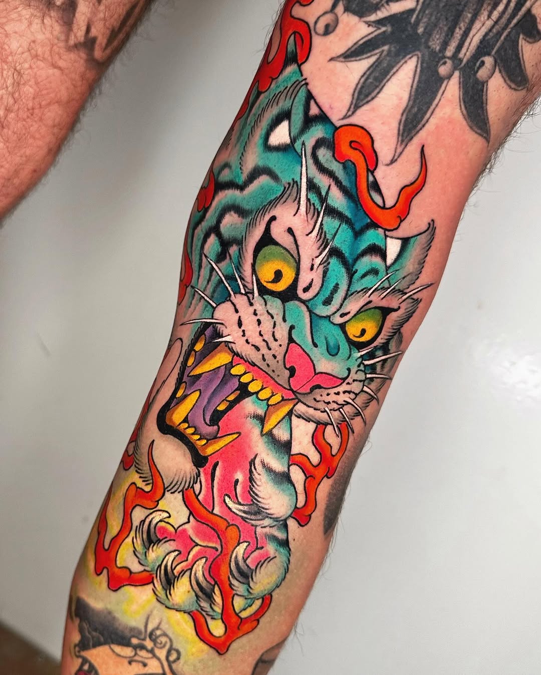 Colorful cat head tattoo on forearm with turquoise face, yellow eyes, red and orange flames surrounding it, and yellow teeth detail.