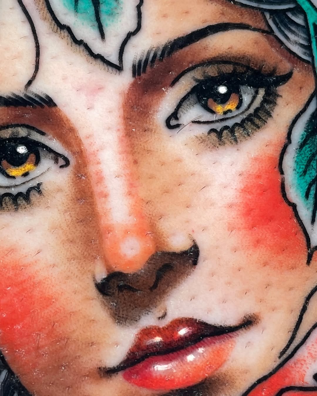 Close-up of a colorful face tattoo featuring golden eyes, red lips, and turquoise hair with black outlines on skin.