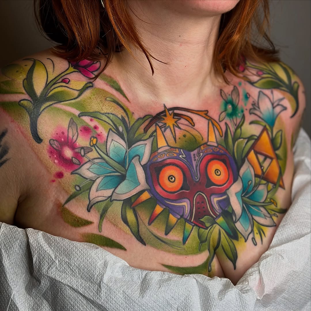 Colorful chest and shoulder tattoo featuring a decorative mask with orange eyes surrounded by flowering branches in yellow, green, teal, and pink tones.