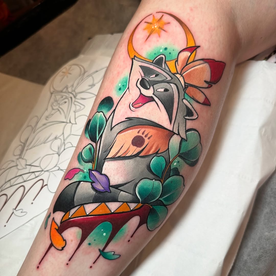 Colorful tattoo on a forearm featuring a stylized face with geometric elements, surrounded by teal leaves and orange celestial symbols against pink skin.