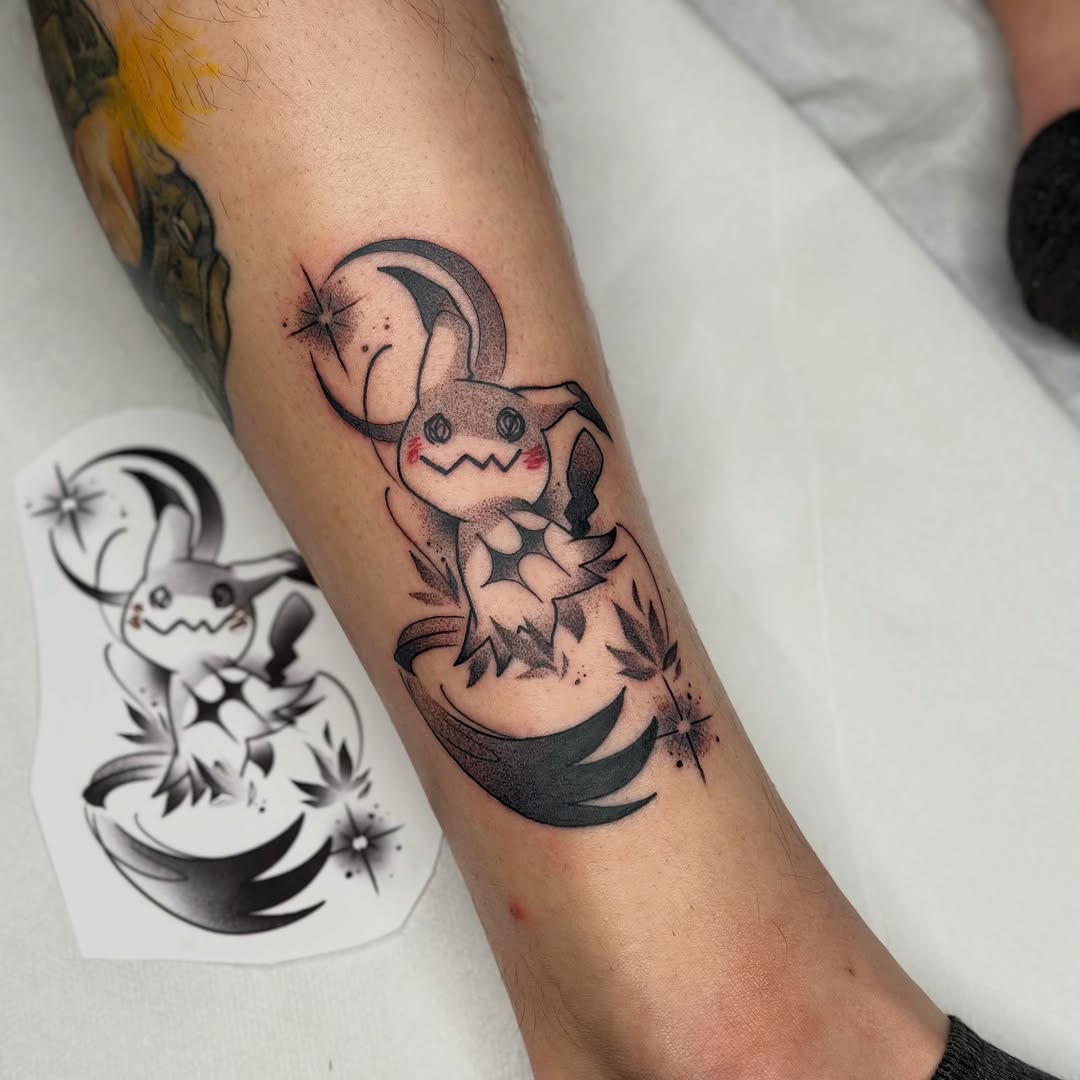 Tattoo featuring a cute character with a crescent moon and star above it, surrounded by decorative elements, on an arm with a reference design visible.