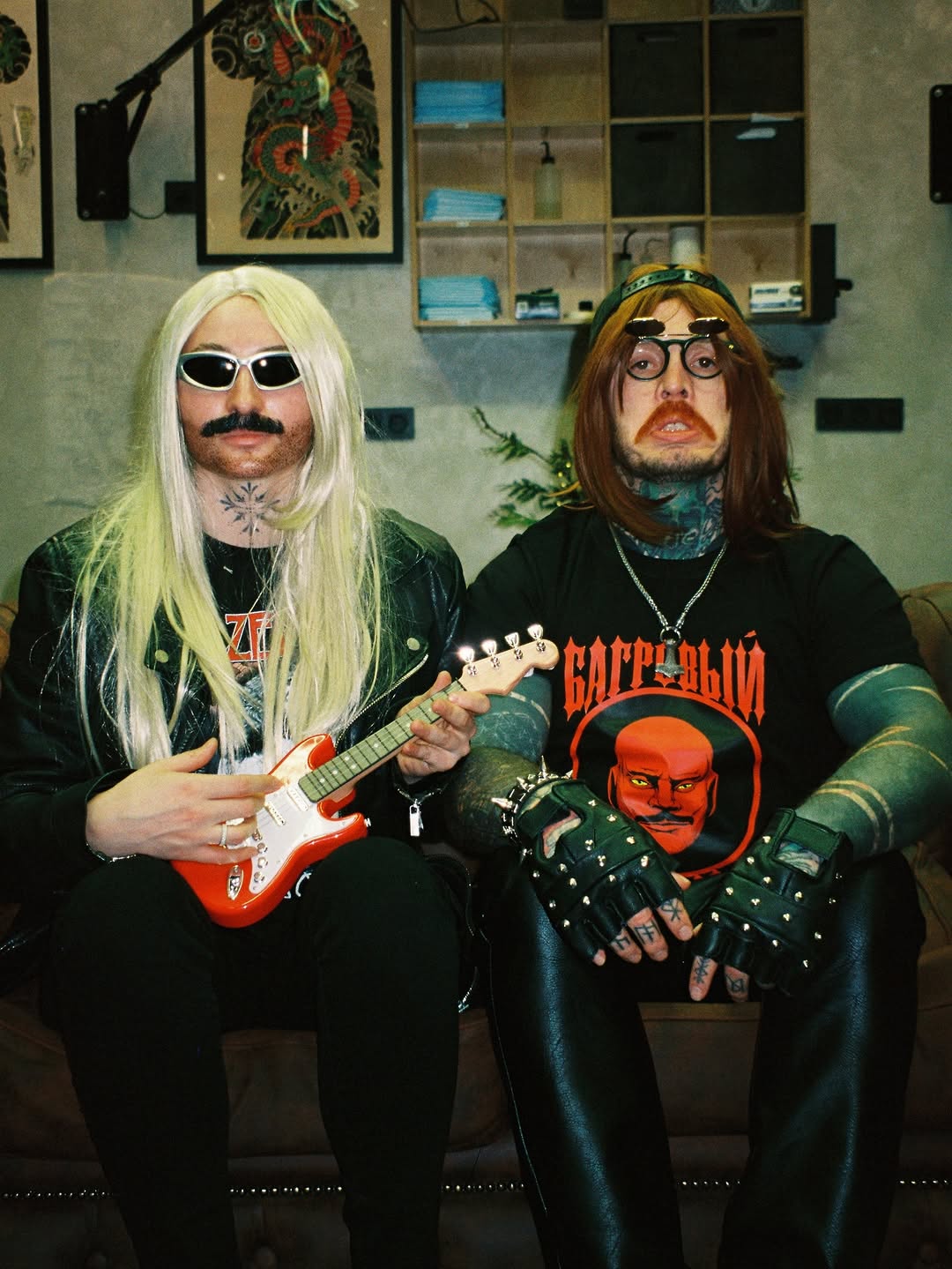 Two musicians with extensive body tattoos pose indoors; left figure displays a chest tattoo, right figure shows neck and hand tattoos in black and colored ink.