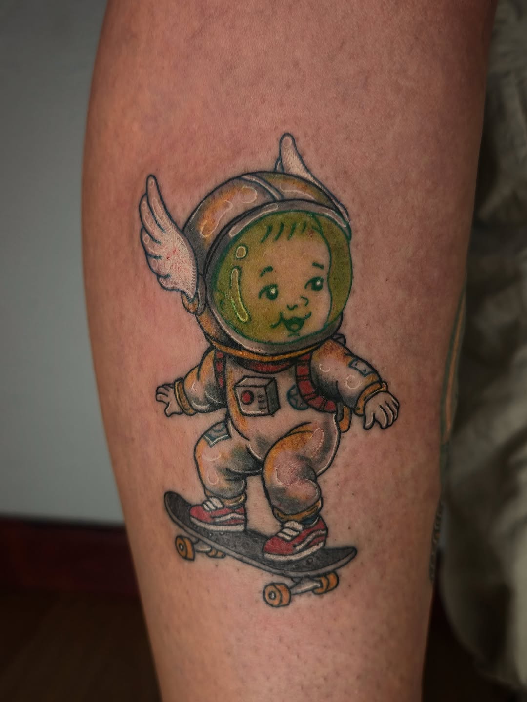 Smiling baby astronaut with wings riding a skateboard, wearing a spacesuit and helmet with green face, tattooed on a calf in tan, green, red, and black colors.