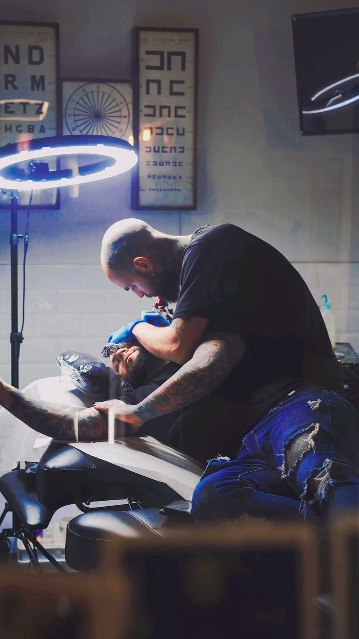 Tattoo artist with visible arm tattoos works on a client's leg using blue lighting equipment in a studio setting.
