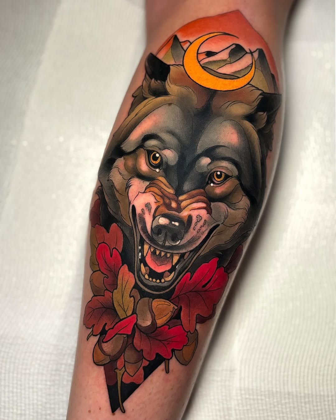 Wolf head with golden eyes and open mouth, topped with crescent moon, surrounded by red and brown autumn leaves on lower leg.