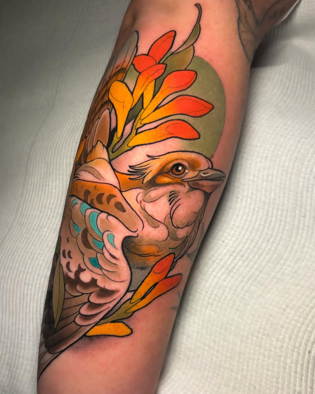Colorful bird tattoo on a calf with orange and red plumage, green foliage, turquoise wing details, and black outline work.