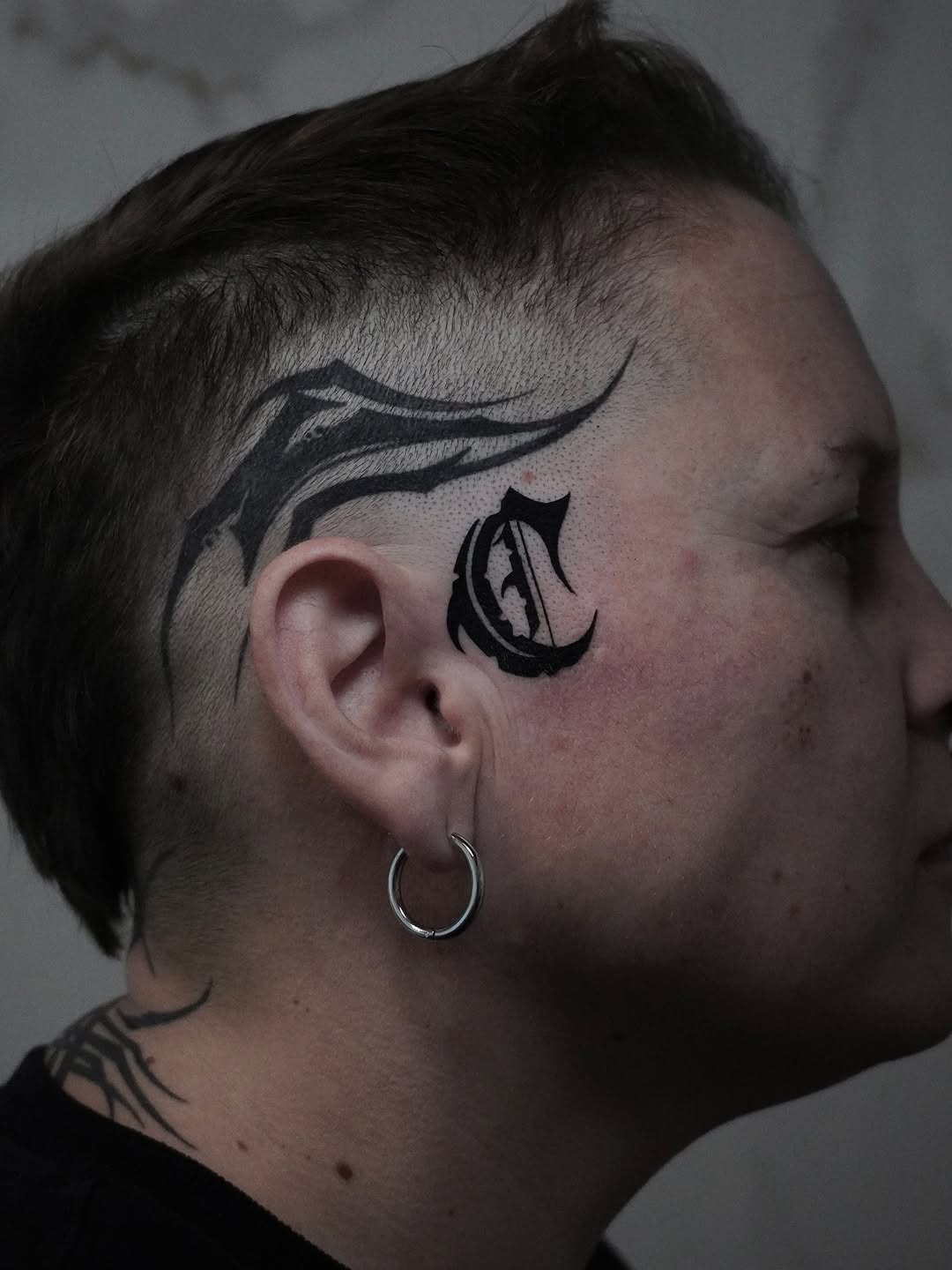 Side head tattoo featuring a curved line design above the ear and a decorative teardrop-shaped symbol with black ink below it.